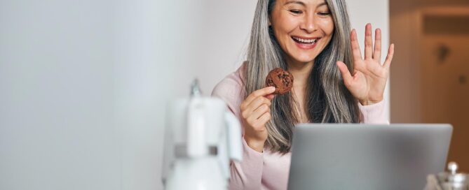 En kvinna sitter bakom en datorskärm och håller en kaka i handen. Vinkar med andra handen mot datorn.